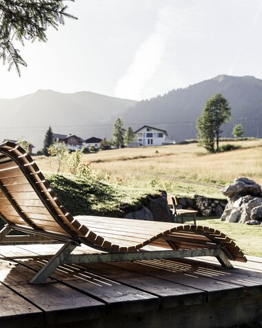 Gemütliche Holzliege auf einem Holzdeck in einer natürlichen, bergigen Umgebung mit Wiesen, Bäumen und Bergen im Hintergrund. | © Kleinwalsertal Tourismus eGen • Fotograf: Oliver Farys