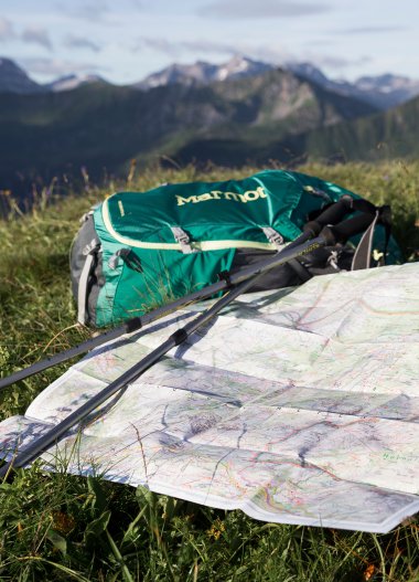 Wanderkarte, Rucksack und Wanderstöcke auf einer Wiese mit Bergpanorama im Hintergrund. | © Kleinwalsertal Tourismus eGen • Fotograf: Oliver Farys
