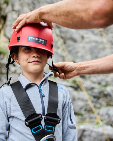 Ein Junge trägt einen Kletterhelm und wird von einem Erwachsenen für eine Bergklettertour vorbereitet. | © Kleinwalsertal Tourismus eGen • Fotograf: Oliver Farys