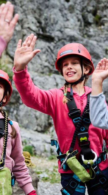 Drei Kinder beim Klettern im Klettergarten, tragen Helme und Kletterausrüstung, lachen und high-fiven sich, Berglandschaft im Hintergrund. | © Kleinwalsertal Tourismus eGen • Fotograf: Oliver Farys