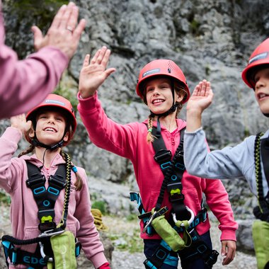 Drei Kinder beim Klettern im Klettergarten, tragen Helme und Kletterausrüstung, lachen und high-fiven sich, Berglandschaft im Hintergrund. | © Kleinwalsertal Tourismus eGen • Fotograf: Oliver Farys