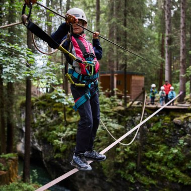 Frau beim Klettern auf einem Seil im Wald bei einem Abenteuerpark, umgeben von Bäumen, mit Helm und Sicherheitsausrüstung. | © Kleinwalsertal Tourismus eGen • Fotograf: Oliver Farys