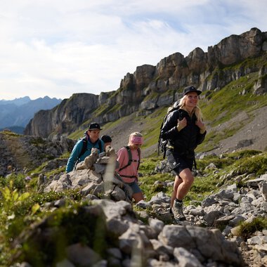 Vier Wanderer klettern auf einem felsigen Bergpfad im Gottesacker im Kleinwalsertal, mit Berggipfeln im Hintergrund an einem sonnigen Tag. | © Kleinwalsertal Tourismus eGen • Fotograf: Oliver Farys