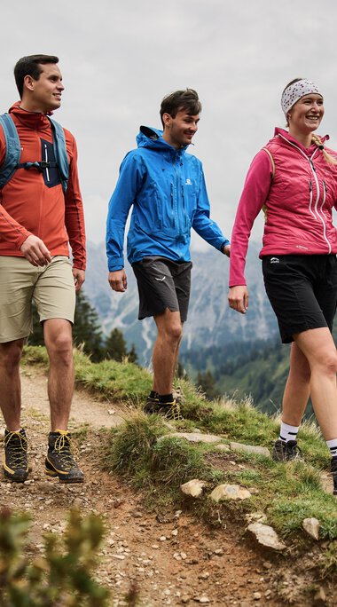 Drei Menschen wandern entlang eines Bergpfads im Kleinwalsertal, umgeben von Bergen und Tannen, bei bewölktem Himmel. | © Kleinwalsertal Tourismus eGen • Fotograf: Oliver Farys