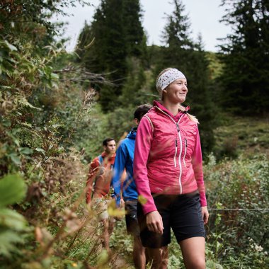 Eine Gruppe von Wanderern, angeführt von einer lachenden Frau in einer pinken Jacke, wandert durch eine grüne, bewaldete Landschaft im Herbst. | © Kleinwalsertal Tourismus eGen • Fotograf: Oliver Farys