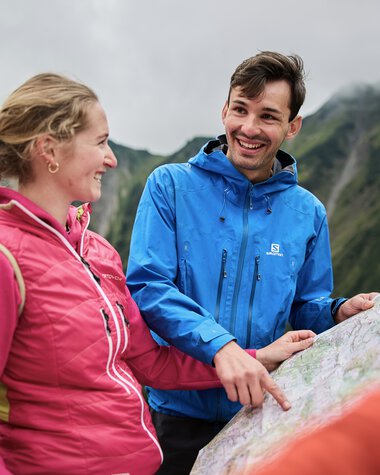 Zwei Wanderer mit Rucksack, lachend und sich unterhaltend während einer Herbstwanderung im Kleinwalsertal, mit Bergpanorama im Hintergrund. | © Kleinwalsertal Tourismus eGen • Fotograf: Oliver Farys