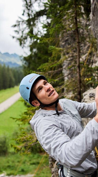 Frau beim Klettern am Klettergarten in den Bergen, umgeben von grünen Wäldern und einer Landschaft mit Weg im Hintergrund. | © Kleinwalsertal Tourismus eGen • Fotograf: Oliver Farys