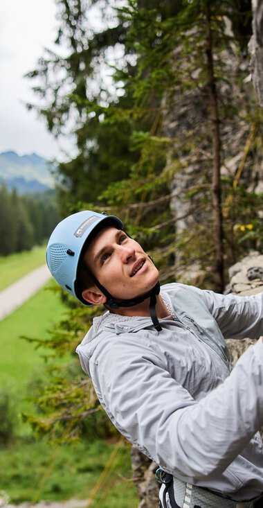 Frau beim Klettern am Klettergarten in den Bergen, umgeben von grünen Wäldern und einer Landschaft mit Weg im Hintergrund. | © Kleinwalsertal Tourismus eGen • Fotograf: Oliver Farys