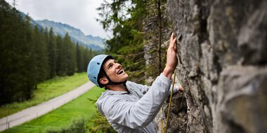 Junge Frau beim Klettern im Klettergarten, trägt Helm und Sicherheitsausrüstung, umgeben von Bergen und Wäldern. | © Kleinwalsertal Tourismus eGen • Fotograf: Oliver Farys