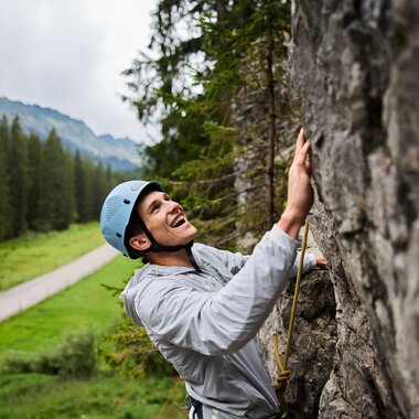Young woman climbing in a rock climbing area, wearing a helmet and safety gear, with mountains and forests in the background. | © Kleinwalsertal Tourismus eGen • Photographer: Oliver Farys