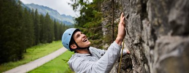 Junge Frau beim Klettern im Klettergarten, trägt Helm und Sicherheitsausrüstung, umgeben von Bergen und Wäldern. | © Kleinwalsertal Tourismus eGen • Fotograf: Oliver Farys