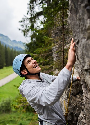 Junge Frau beim Klettern im Klettergarten, trägt Helm und Sicherheitsausrüstung, umgeben von Bergen und Wäldern. | © Kleinwalsertal Tourismus eGen • Fotograf: Oliver Farys