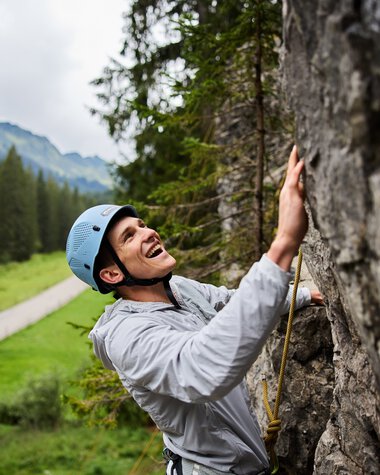 Junge Frau beim Klettern im Klettergarten, trägt Helm und Sicherheitsausrüstung, umgeben von Bergen und Wäldern. | © Kleinwalsertal Tourismus eGen • Fotograf: Oliver Farys