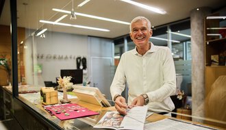 Mann im Büro hinter dem Kundenservice-Schalter, lächelnd, mit Unterlagen und Kasse. | © Kleinwalsertal Tourismus eGen • Fotograf: Oliver Farys