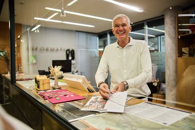 Mann im Büro hinter dem Kundenservice-Schalter, lächelnd, mit Unterlagen und Kasse. | © Kleinwalsertal Tourismus eGen • Fotograf: Oliver Farys