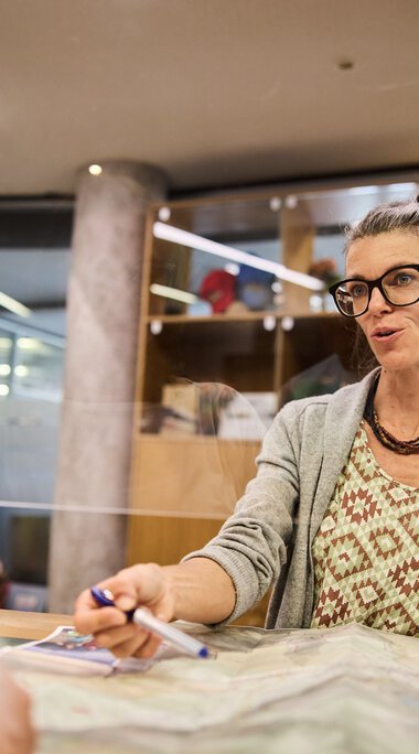 Frau mit Brille und lockigem Haar sitzt an einem Tisch in einem Kundenservice-Büro, spricht freundlich und hat Papiere vor sich. | © Kleinwalsertal Tourismus eGen • Fotograf: Oliver Farys