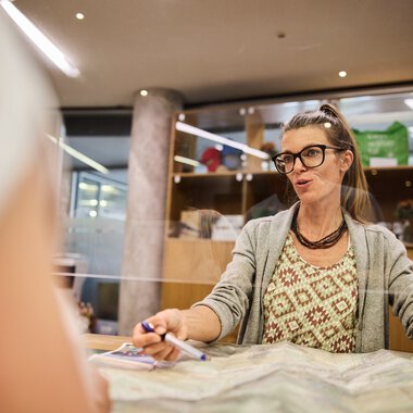 Woman with glasses and curly hair sitting at a desk in a customer service office, speaking friendly, with papers in front of her. | © Kleinwalsertal Tourismus eGen • Photographer: Oliver Farys