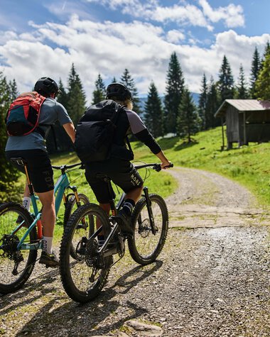 Zwei Mountainbiker auf einem Waldpfad im Kleinwalsertal, umgeben von Bäumen und einer kleinen Hütte im Hintergrund, sonniger Himmel. | © Kleinwalsertal Tourismus eGen • Fotograf: Oliver Farys