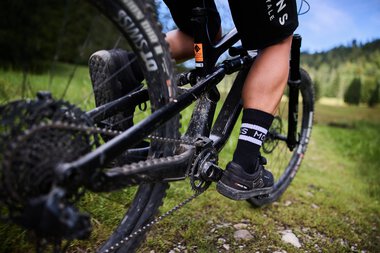 Nahaufnahme eines Mountainbikers, der auf einem Singletrail im Kleinwalsertal fährt, mit Fokus auf die Pedale und die Kette. | © Kleinwalsertal Tourismus eGen • Fotograf: Oliver Farys