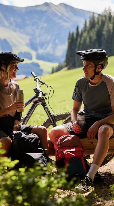 Zwei Mountainbike-Fahrer in Helm, sitzend auf einer Bank im Grünen, unterhalb eines Baumes, mit Mountainbikes im Hintergrund bei Sommer im Kleinwalsertal. | © Kleinwalsertal Tourismus eGen • Fotograf: Oliver Farys