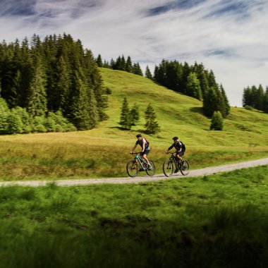 Zwei Radfahrer auf einem Waldweg im Kleinwalsertal, umgeben von grünen Hügeln und Wäldern, bei schönem Wetter. | © Kleinwalsertal Tourismus eGen • Fotograf: Oliver Farys