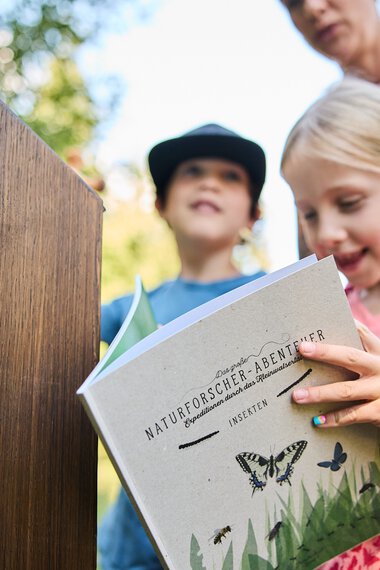 Ein Mädchen liest ein Buch über Insekten im Naturforscherabenteuer in Kleinwalsertal, begleitet von Erwachsenen und einem anderen Kind im Hintergrund. | © Kleinwalsertal Tourismus eGen • Fotograf: Oliver Farys