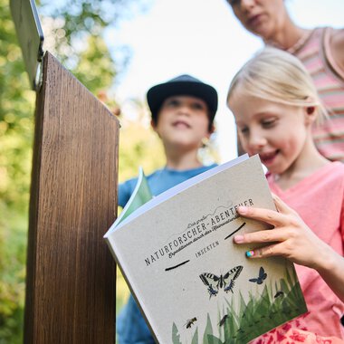 Ein Mädchen liest ein Buch über Insekten im Naturforscherabenteuer in Kleinwalsertal, begleitet von Erwachsenen und einem anderen Kind im Hintergrund. | © Kleinwalsertal Tourismus eGen • Fotograf: Oliver Farys