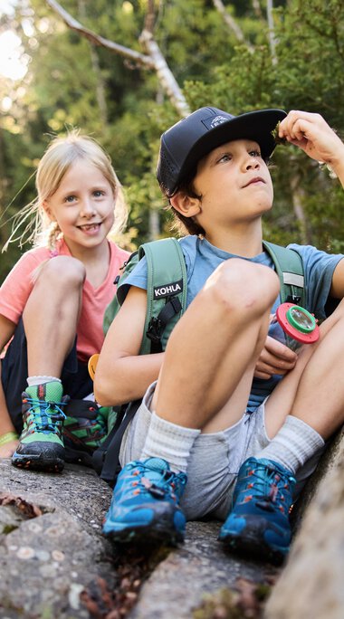 Zwei Kinder auf einem Felsen in einem Wald, eines schaut nach oben, das andere lächelt, Umgeben von Bäumen und Natur. | © Kleinwalsertal Tourismus eGen • Fotograf: Oliver Farys