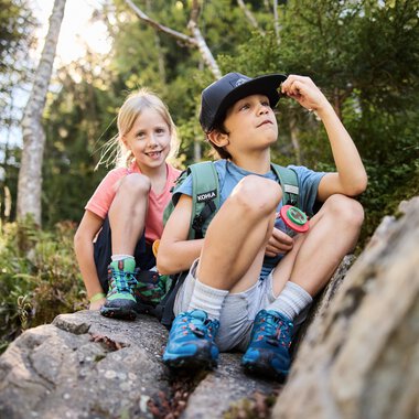 Zwei Kinder auf einem Felsen in einem Wald, eines schaut nach oben, das andere lächelt, Umgeben von Bäumen und Natur. | © Kleinwalsertal Tourismus eGen • Fotograf: Oliver Farys