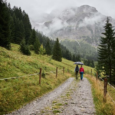 Wanderer mit Regenschirm auf einem Kiesweg im bergigen, nebligen Gebiet mit Tannenbäumen und Bergen im Hintergrund. | © Kleinwalsertal Tourismus eGen • Fotograf: Oliver Farys