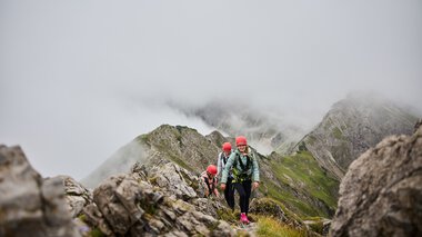 © Kleinwalsertal Tourismus eGen • Fotograf: Oliver Farys Menschen beim Klettersteig in den Bergen, umgeben von Wolken und Felsen, mit Bergschule Walter Walser-Logo im Bild. | © Kleinwalsertal Tourismus eGen • Fotograf: Oliver Farys