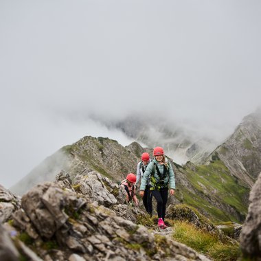 Menschen beim Klettersteig in den Bergen, umgeben von Wolken und Felsen, mit Bergschule Walter Walser-Logo im Bild. | © Kleinwalsertal Tourismus eGen • Fotograf: Oliver Farys