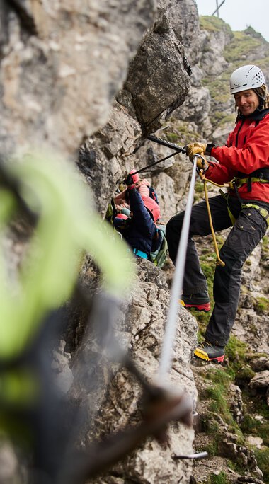Frauen beim Klettersteig in den Bergen, Sicherungsgurte tragen, Naturkulisse im Hintergrund.