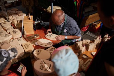Ein älterer Mann beim Signieren oder Bemalen von Holzartikeln auf einem Marktstand im Kleinwalsertal. | © Kleinwalsertal Tourismus eGen • Fotograf: Oliver Farys