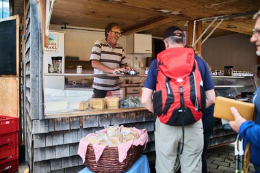 Verkäuferin beim Wochenmarkt im Kleinwalsertal im Sommer 2023, die frisches Gemüse und Käse anbietet, während Kunden mit Rucksack einkaufen. | © Kleinwalsertal Tourismus eGen • Fotograf: Oliver Farys