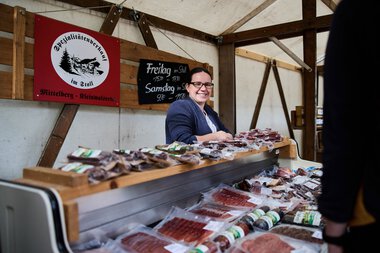 Frau verkauft Fleischwaren am Walser Wochenmarkt im Kleinwalsertal, im Hintergrund ein Schild mit Wochenmarktzeiten. | © Kleinwalsertal Tourismus eGen • Fotograf: Oliver Farys