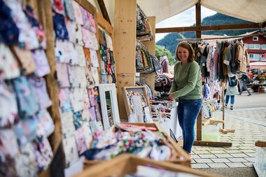 Frau verkauft handgemachte Stoffe auf einem Wochenmarkt im Kleinwalsertal, umgeben von bunten Textilien und einer malerischen Bergkulisse. | © Kleinwalsertal Tourismus eGen • Fotograf: Oliver Farys