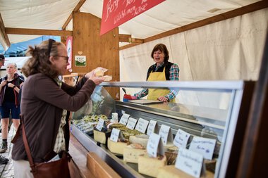Fotografie von einem Verkaufsstand bei einem Wochenmarkt im Kleinwalsertal, bei dem eine Verkäuferin mit Kunden Käse verkauft. | © Kleinwalsertal Tourismus eGen • Fotograf: Oliver Farys