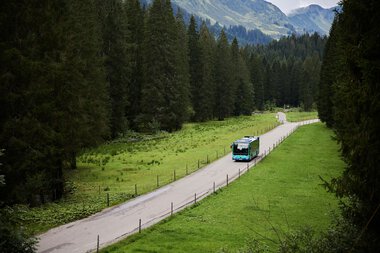 Ein Bus fährt auf einer kleinen Landschaftstraße durch eine grüne Wiesen- und Waldregion in den Bergen, mit Bergen im Hintergrund. | © Kleinwalsertal Tourismus eGen • Fotograf: Oliver Farys
