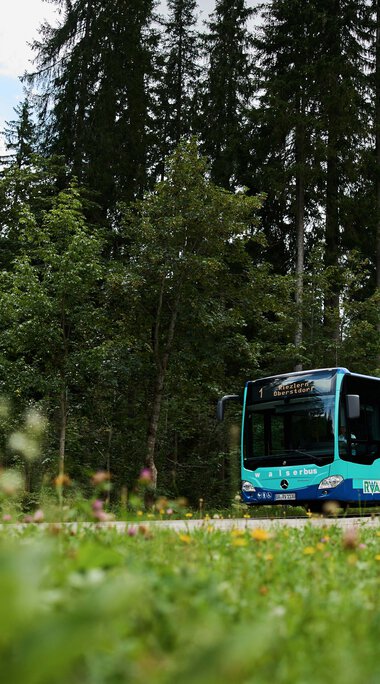 Ein moderner Bus fährt durch eine grüne, von Wald umgebene Landschaft bei schönem Wetter. | © Kleinwalsertal Tourismus eGen • Fotograf: Oliver Farys