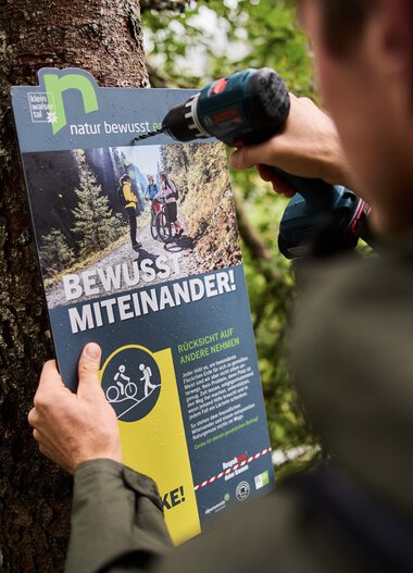 Ein Ranger befestigt ein Informationsschild im Wald, um Naturschutzmaßnahmen und Wissensvermittlung zu fördern. | © Kleinwalsertal Tourismus eGen • Fotograf: Oliver Farys