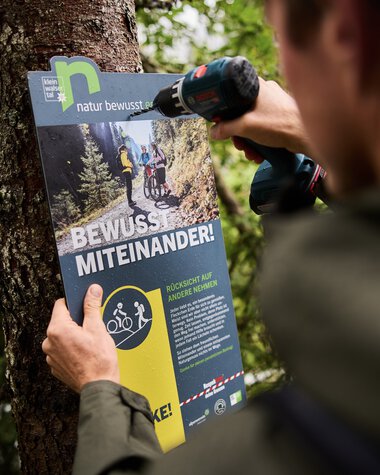 Ein Ranger befestigt ein Informationsschild im Wald, um Naturschutzmaßnahmen und Wissensvermittlung zu fördern. | © Kleinwalsertal Tourismus eGen • Fotograf: Oliver Farys