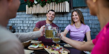 Four friends cheers with beer mugs during an outdoor meal, with a variety of snacks on the table, in front of a traditional alpine building decorated with flower boxes. | © Kleinwalsertal Tourismus eGen • Photographer: Oliver Farys
