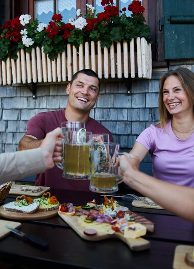 Vier Freunde stoßen beim Essen im Freien an, mit einem Berg von Snacks und Bier, vor einem traditionellen Alpengebäude mit Blumentopf. | © Kleinwalsertal Tourismus eGen • Fotograf: Oliver Farys