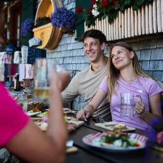 Zwei lachende Personen sitzen bei einem Sommeressen im Freien an einem Tisch vor einer alpenländischen Hütte, unter einer Fensterbank mit Blumen. | © Kleinwalsertal Tourismus eGen • Fotograf: Oliver Farys