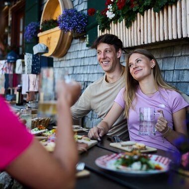 Zwei lachende Personen sitzen bei einem Sommeressen im Freien an einem Tisch vor einer alpenländischen Hütte, unter einer Fensterbank mit Blumen. | © Kleinwalsertal Tourismus eGen • Fotograf: Oliver Farys