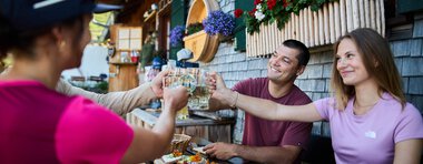 Menschen beim Sommer-Outdooressen auf einer Terrasse mit Holzhäuschen und bunten Blumen, Toasts und Snacks werden ausgetauscht. | © Kleinwalsertal Tourismus eGen • Fotograf: Oliver Farys