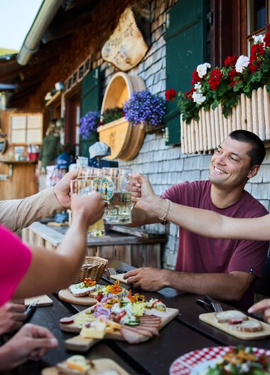 Menschen beim Sommer-Outdooressen auf einer Terrasse mit Holzhäuschen und bunten Blumen, Toasts und Snacks werden ausgetauscht. | © Kleinwalsertal Tourismus eGen • Fotograf: Oliver Farys