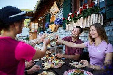 Menschen beim Sommer-Outdooressen auf einer Terrasse mit Holzhäuschen und bunten Blumen, Toasts und Snacks werden ausgetauscht. | © Kleinwalsertal Tourismus eGen • Fotograf: Oliver Farys