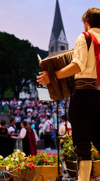 Musiker auf einer Bühne beim Alpenzauber-Festival im Kleinwalsertal, spielt Gitarre während die Menge im Hintergrund zuschaut, mit Bergkulisse und Kirche im Hintergrund. | © Kleinwalsertal Tourismus eGen • Fotograf: Oliver Farys
